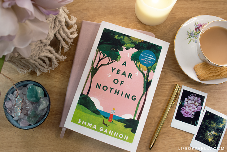 a photo of the books cover with various decorative items around it, a cup of tea, a journal and pen, photos, flowers and crystals